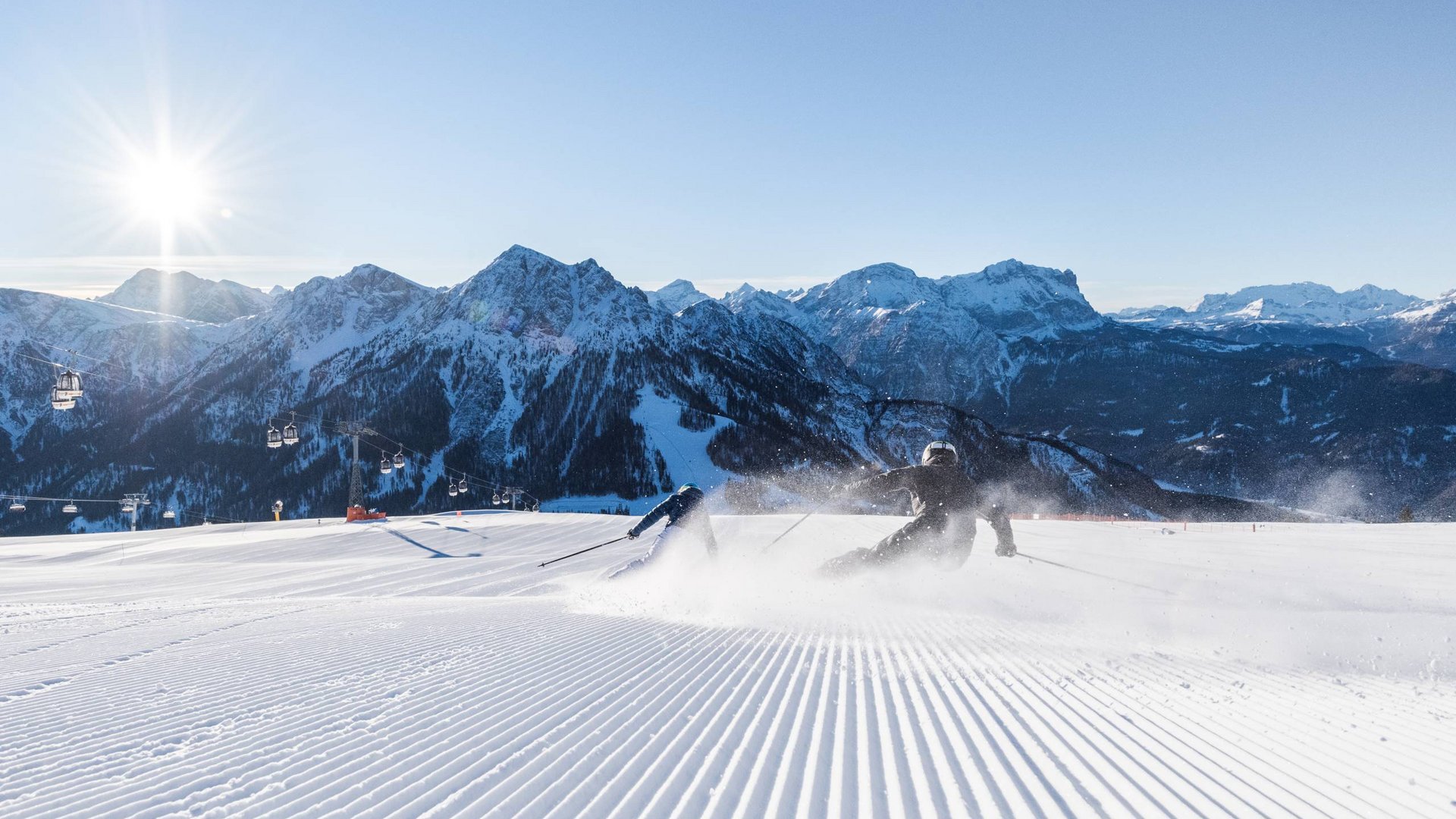 Zwei Skifahrer fahren auf frisch gespurtem Schnee vor Bergpanorama bei Sonnenschein